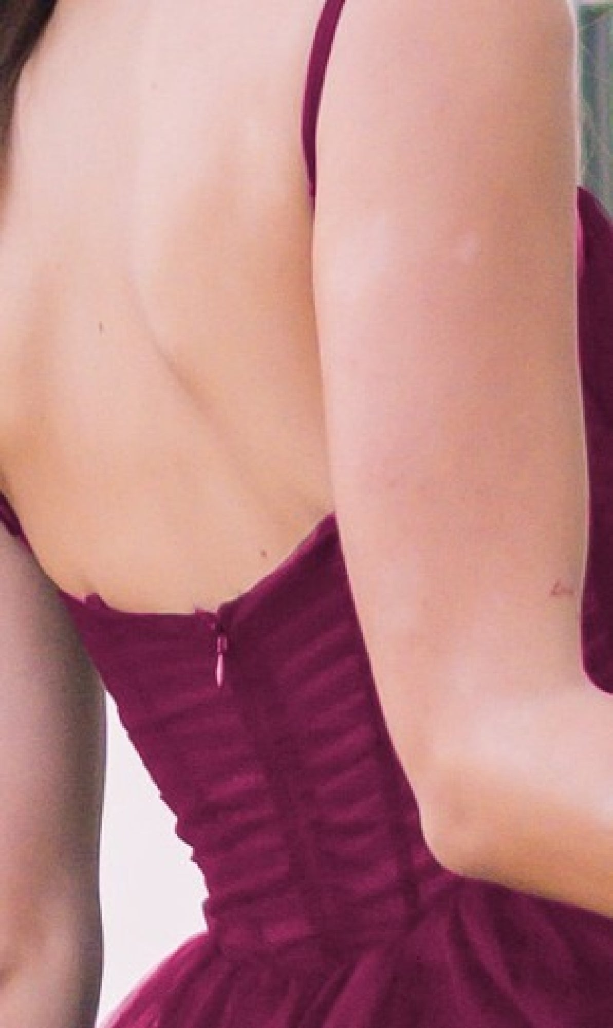A person seen from behind wears the Sheer-Corset Long Prom Ball Gown: Yasmin by Velvi, a sleeveless burgundy dress with a sheer corset bodice and low back, revealing their upper back and part of one arm.